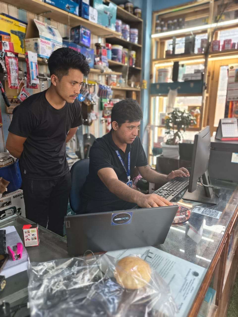 aBit consultant setting up aBitPOS on a laptop at the Kezang Enterprise counter while a staff member watches, hardware shelves behind